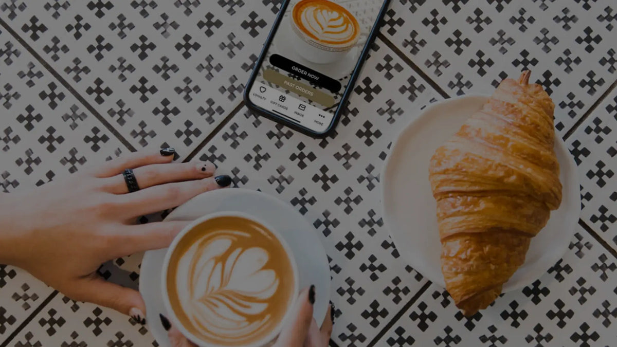 Person holding a cup of latte with foam art beside a croissant and a smartphone displaying the same coffee image on a patterned table