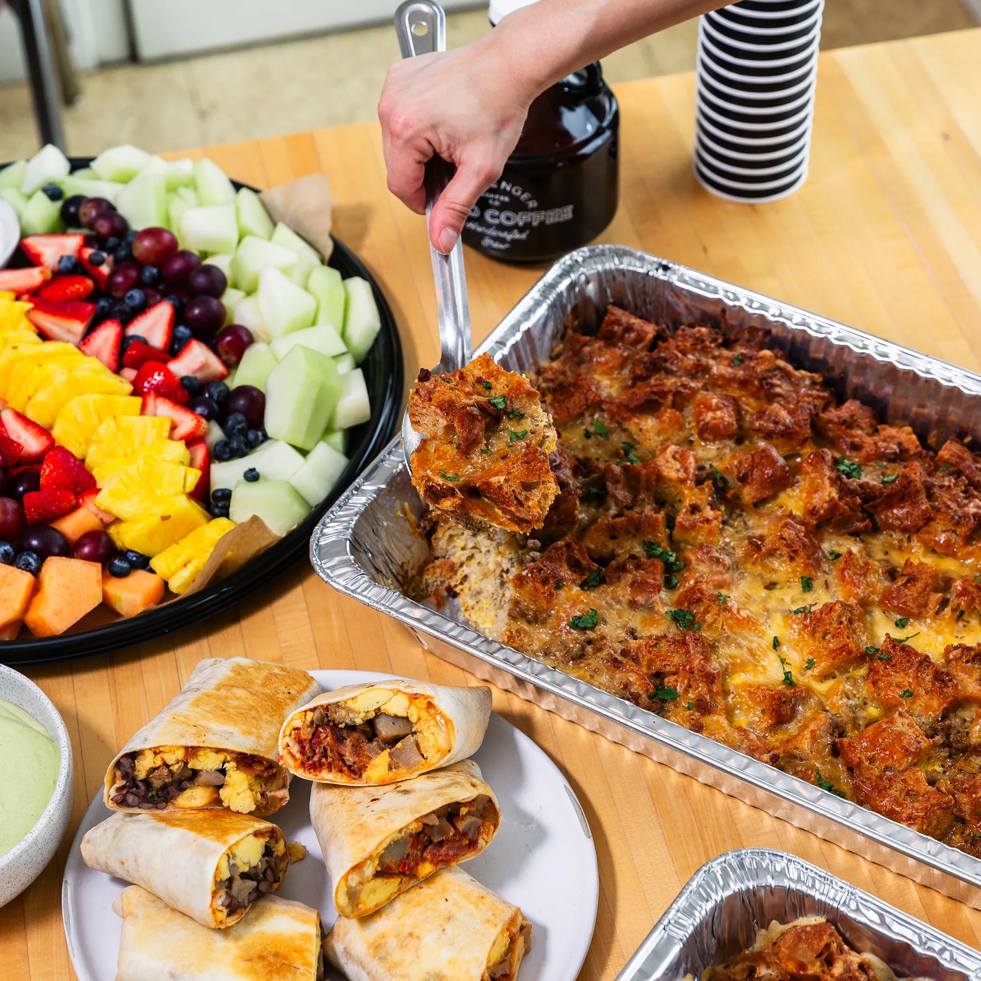 A fruit tray, messenger cold brew coffee, burrito and casserole catering spread on the table. A hand is reaching and scooping out the breakfast casserole.