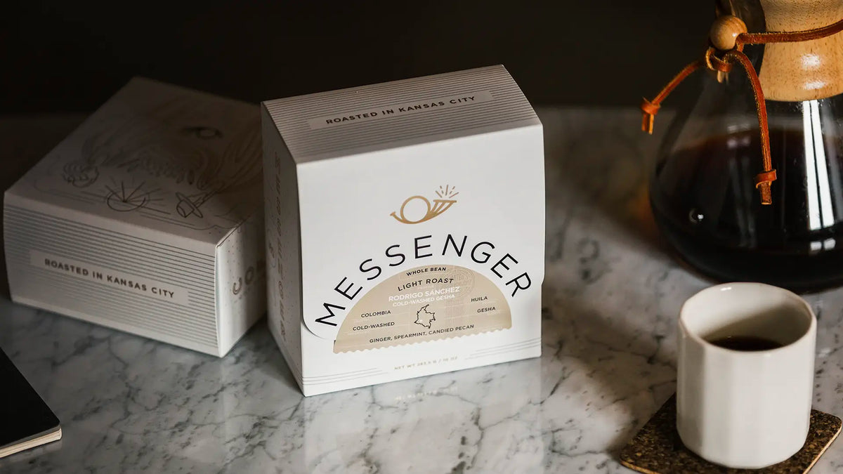 Light roast Messenger coffee package on marble table near cup and coffee brewer in dim light