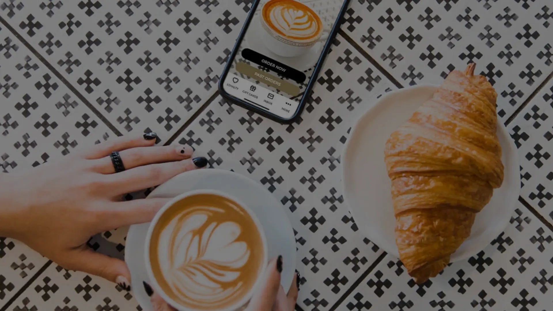 Person holding a cup of latte with foam art beside a croissant and a smartphone displaying the same coffee image on a patterned table